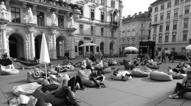 Hier sieht man Menschen auf dem Hauptplatz in Graz. Sie sitzen in Sitzsäcken und Sonnenstühlen, Z.T unter Schirmen, ruhen und lesen.