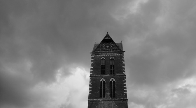 Hier ist der Turm einer Kirche in Wismar zu sehen, vor einem sehr bewölkten Himmel