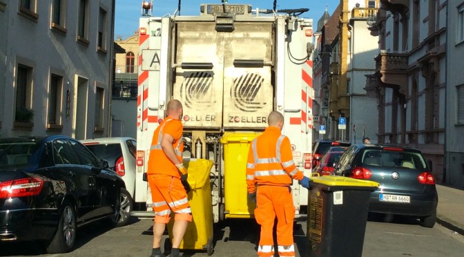 Hier ist ein Bild eines Müllwagens von hinten zu sehen. Zwei Mitarbeiter der Stadt sind gerade dabei, die gelben Tonnen in die Vorrichtungen einzuhängen.