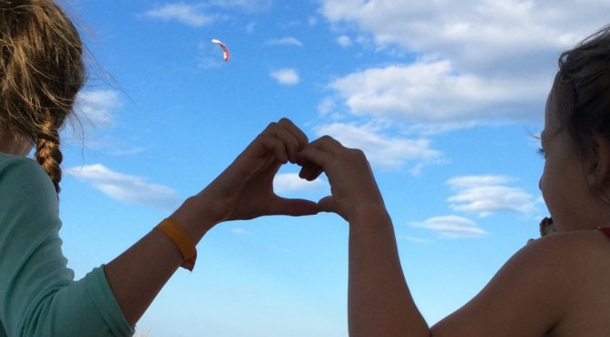 Hier sieht man zwei Kinder, die zusammen mit ihren Händen ein Herz formen, im Hintergrund der blaue Himmel